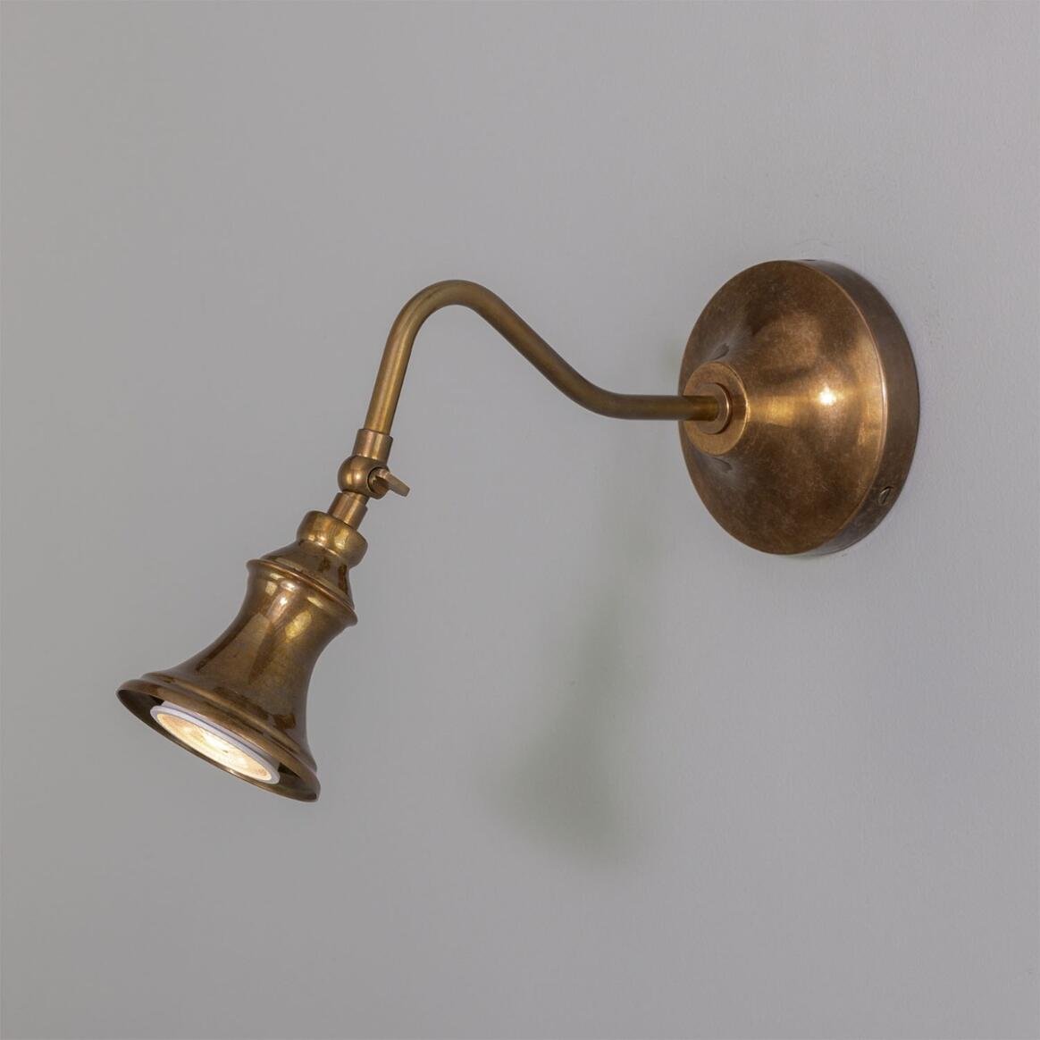 The Adjustable Bell Sconce, featuring a round base, curved arm, and bell-shaped shade in antique brass, is mounted on a plain light-colored wall—its classic design brings timeless elegance to any space.