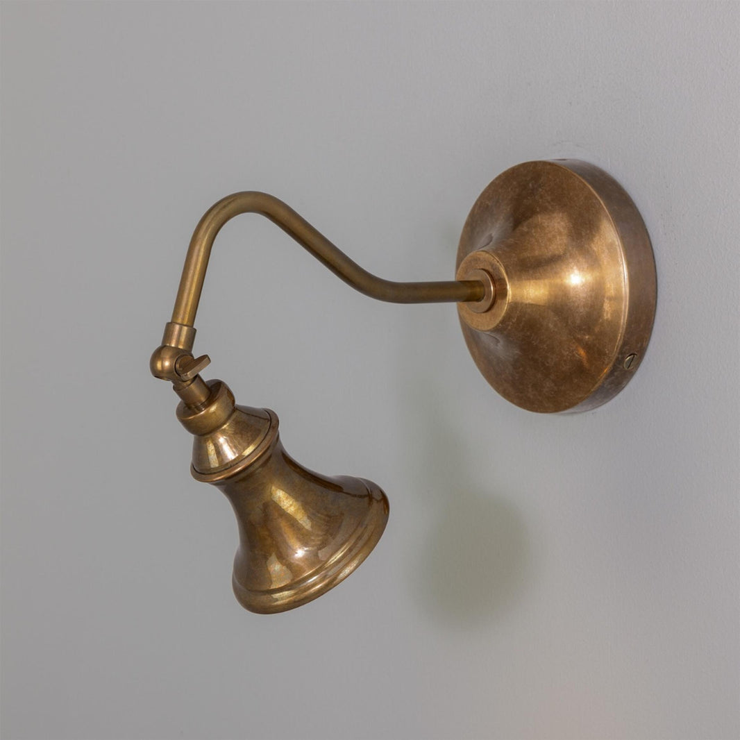 The Adjustable Bell Sconce, crafted in antique brass, features a classic round base and an angled arm supporting a downward-facing bell-shaped shade, shown here against a plain light gray wall.