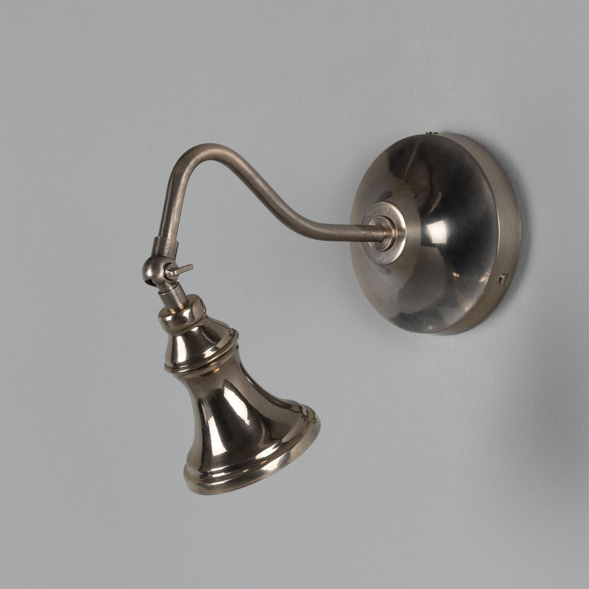 The Adjustable Bell Sconce showcases a timeless metallic design, featuring a round base and an adjustable arm that supports its signature bell-shaped light fixture in antique brass, beautifully mounted on a plain grey wall.