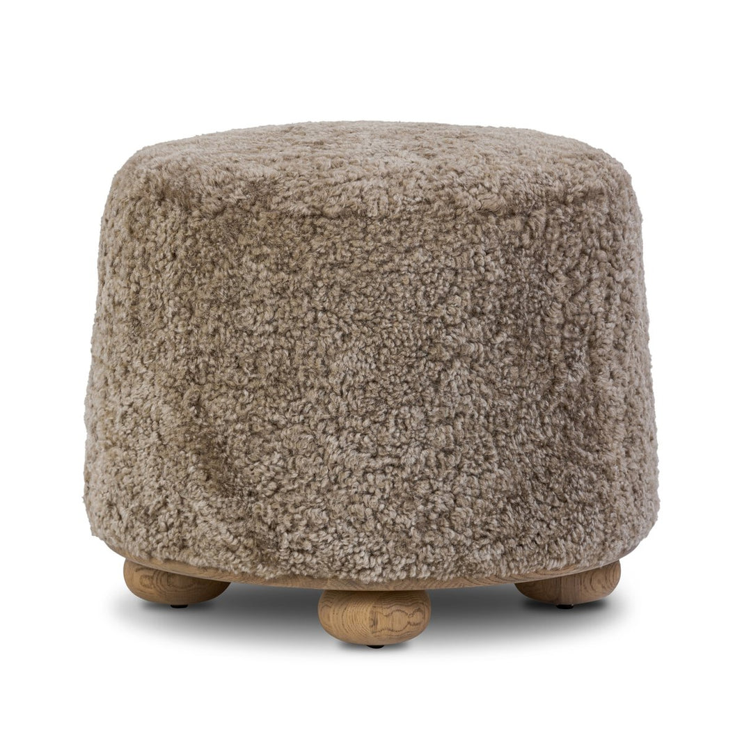 The Taupe Shierling Ottoman features a round design, soft textured beige upholstery, and four short wooden legs.
