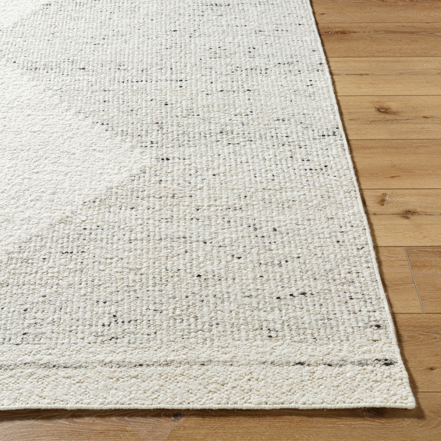 A close-up shows the Large Diamond Wool Rug - 8 x 10, featuring a cream-colored, hand-woven 100% wool texture with dark speckles, placed on a light wood floor and highlighting its detailed craftsmanship.