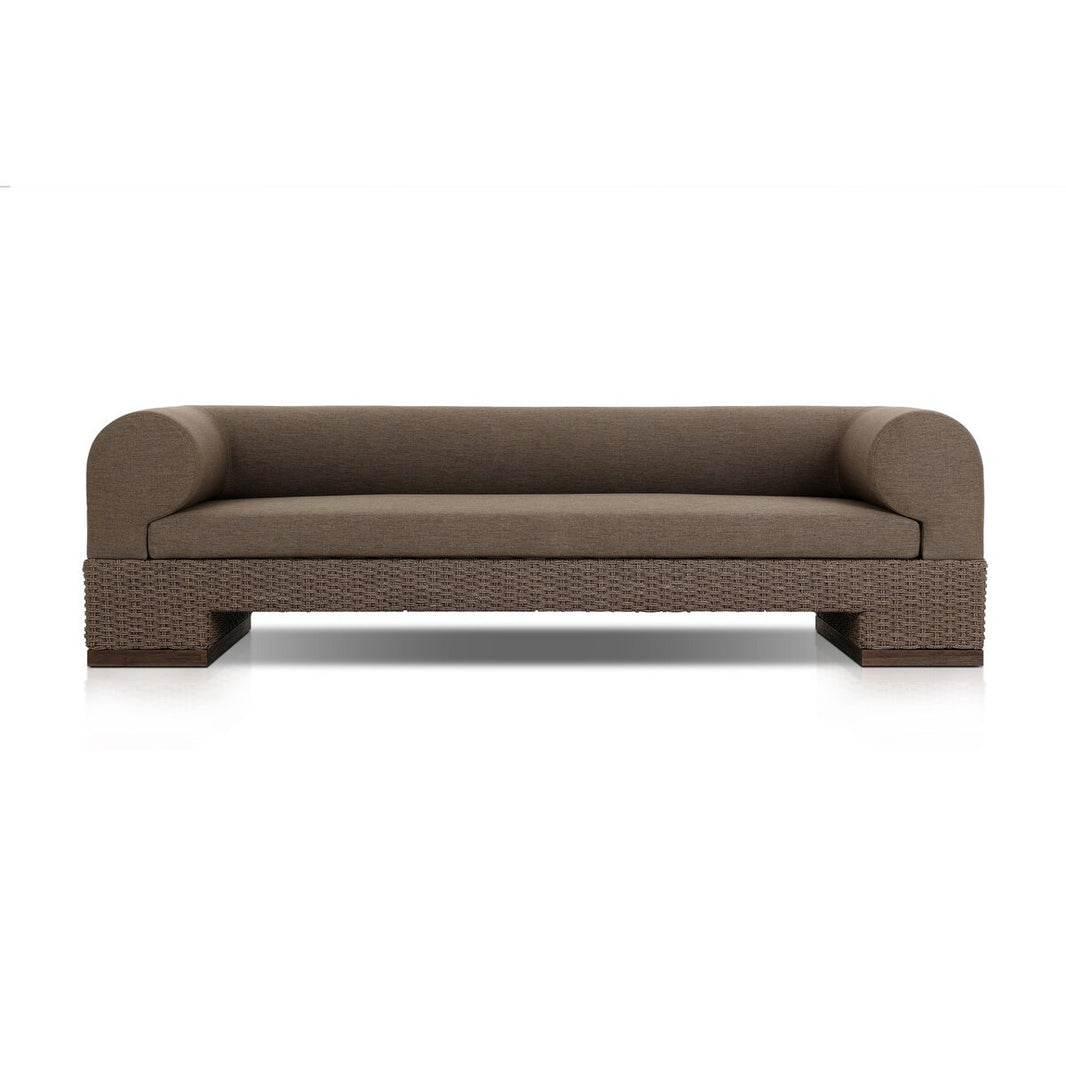 The Wicker Base Outdoor Sofa features a modern brown design with a low back and wide, rounded armrests, a woven wicker base, and a minimalist look—ideal for stylish outdoor seating against any backdrop.