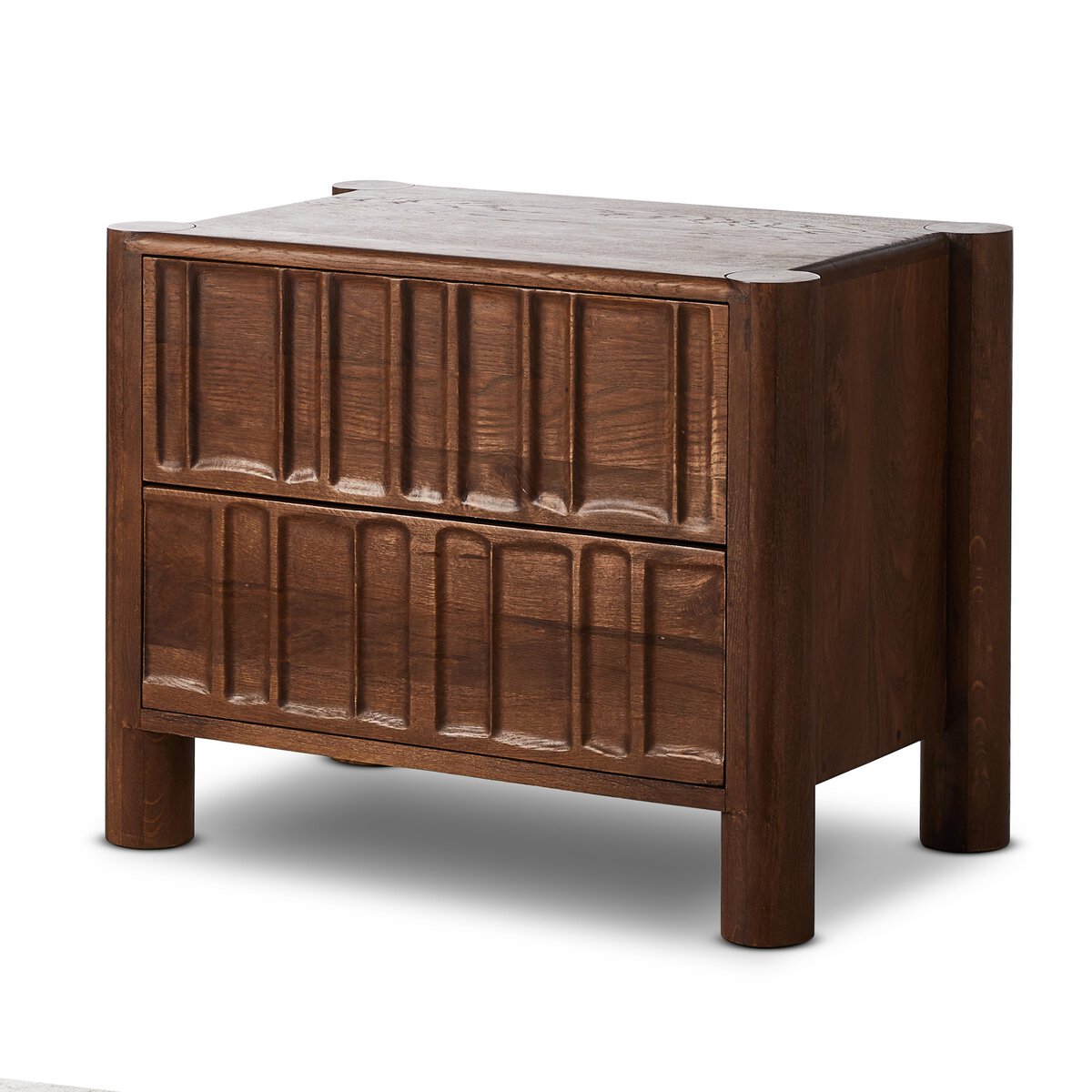 The Primitive Nightstand is a wooden piece made from cocoa-finished oak, featuring two drawers with organic carved grooves on the front and sturdy cylindrical corner legs for a dark, rich look.