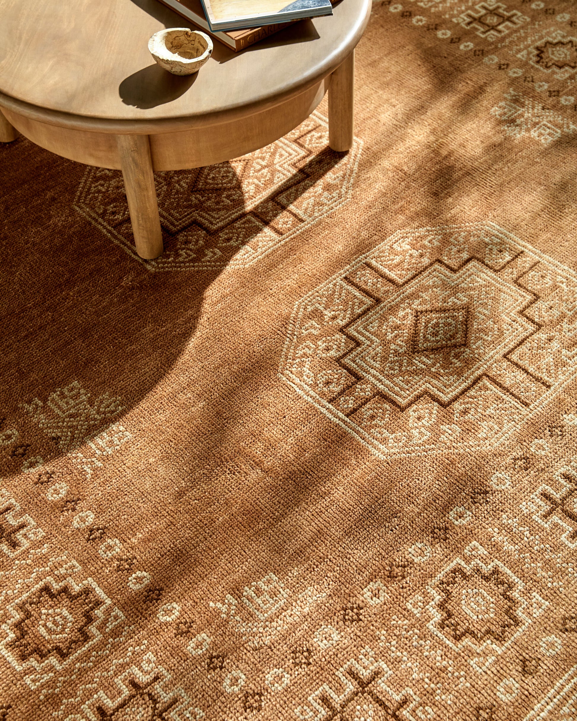 A round wooden coffee table with books and a small bowl sits on a 3' x 10' Burnt Orange Tribal Runner Rug, its geometric patterns highlighted by natural sunlight.