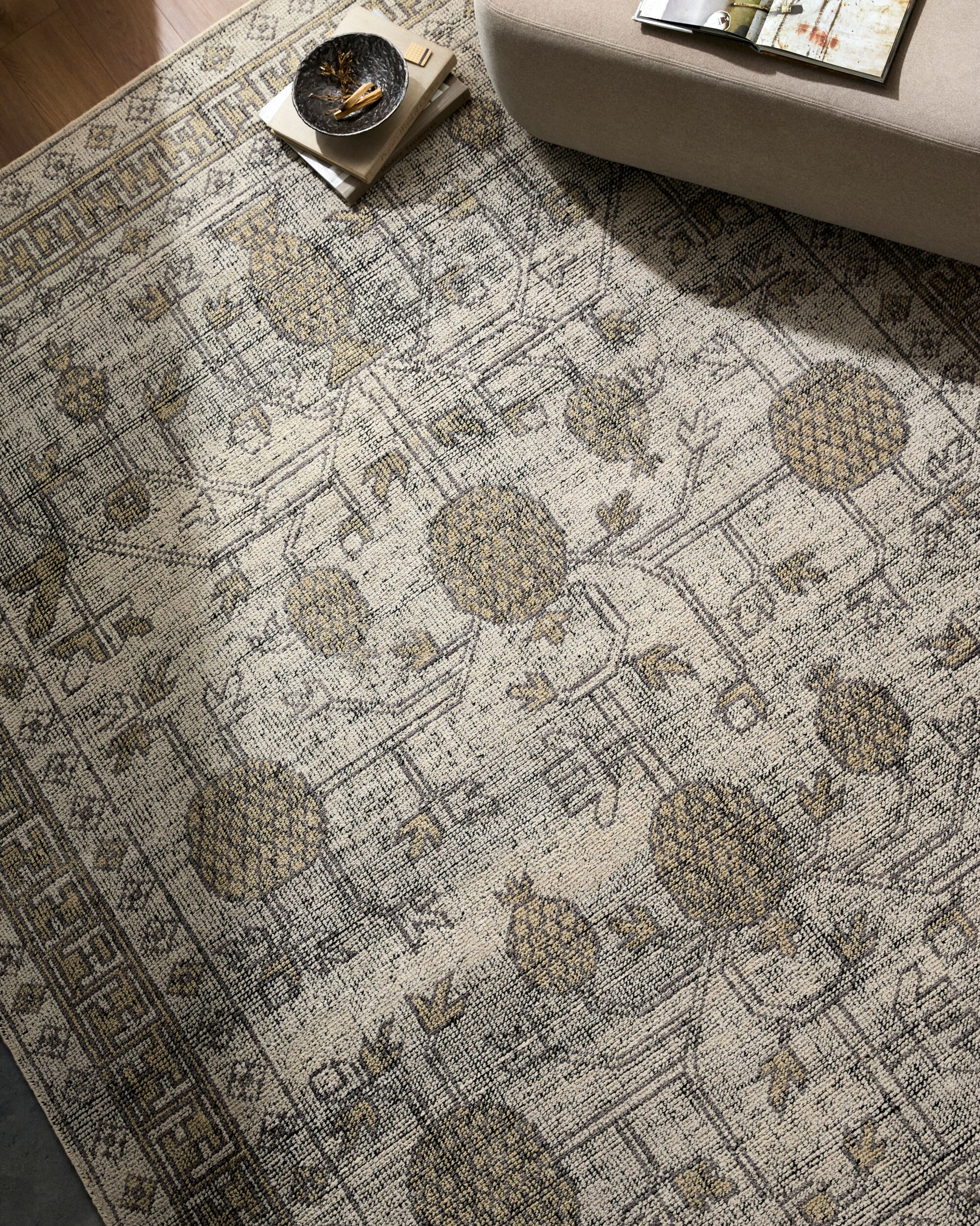 The Patterned 100% Wool Rug - 8'x10' with beige and brown floral and geometric designs sits beside a light beige sofa, holding a stack of books with a bowl on top. A magazine is placed on the sofa. Various sizes available.