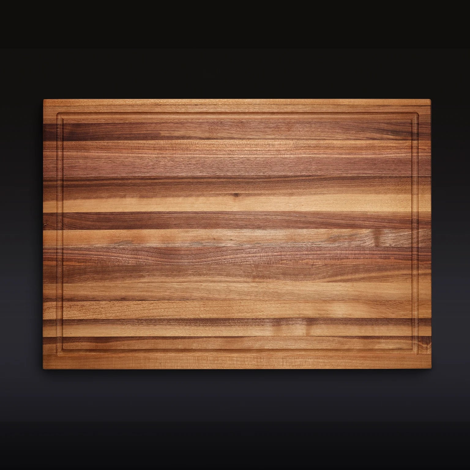 The Hexclad Cutting Board is a rectangular wood board with striking light and dark grain patterns and a shallow groove along the edge, shown against a dark background.